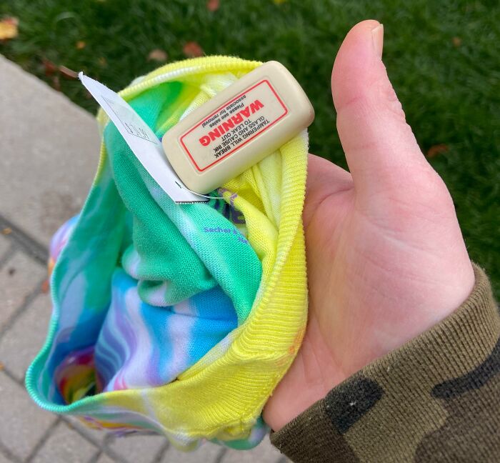Hand holding a tie-dye Halloween costume with a warning tag and security alarm attached, a Halloween fail example.