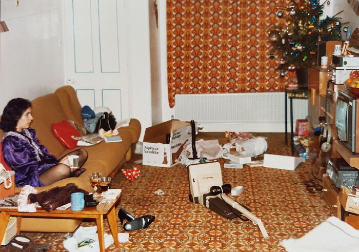 Woman in a purple dress sitting on a couch in a cluttered living room with vintage 1980s decor and patterned carpet.