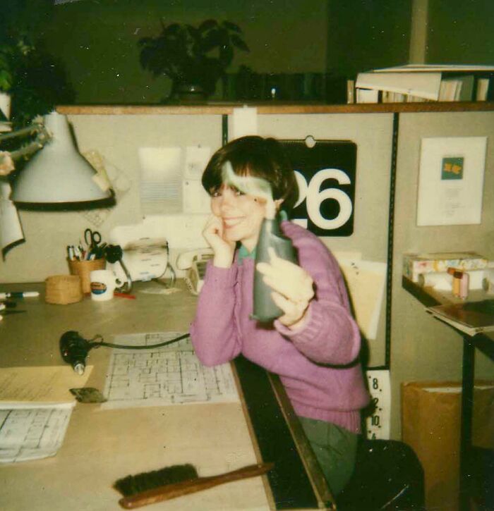 Woman in a purple sweater holding a smoking object at her office desk in a wild and wonderful 1980s setting
