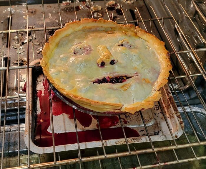 Burnt Halloween pie with a face inside an oven, a funny Halloween fail spilling filling onto a tray below.