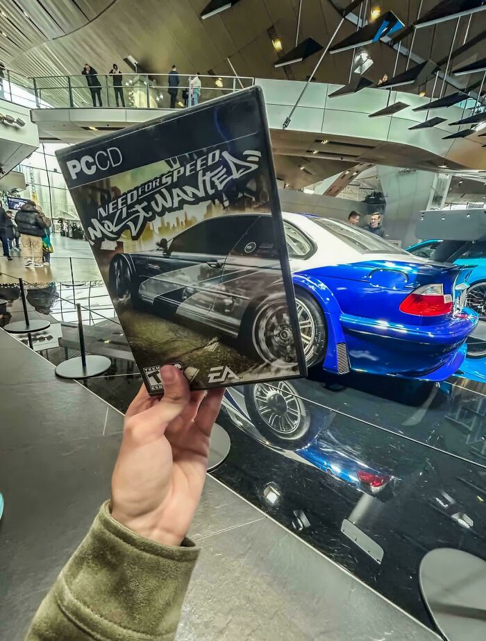 Hand holding Need for Speed Most Wanted game case in front of a blue sports car at a car exhibition, the picture goes hard.