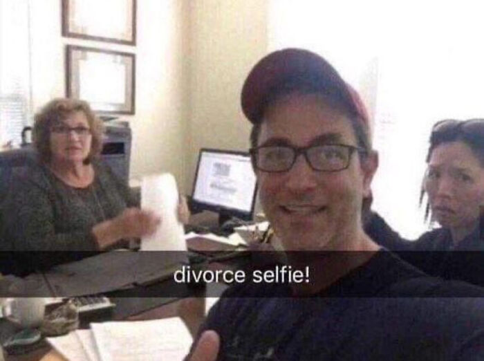 Man wearing glasses and a cap taking a funny selfie in an office during divorce paperwork with two serious women nearby.