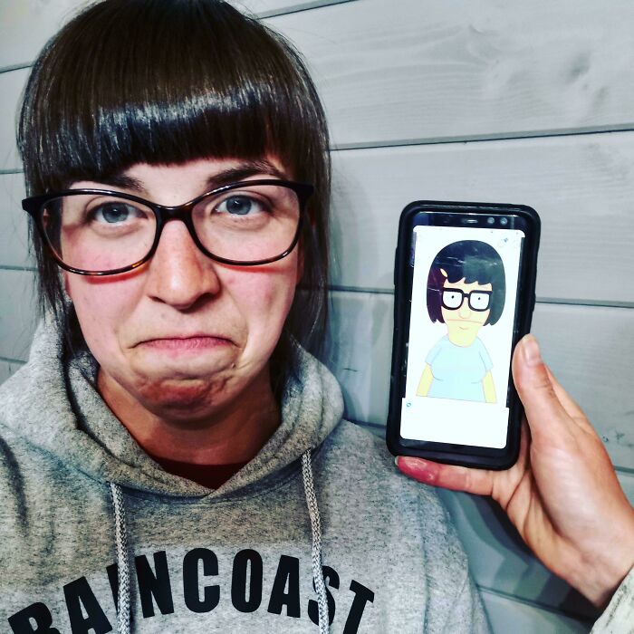 Woman with tragic hair accident making a funny face next to a phone showing a cartoon character with similar hairstyle and glasses.