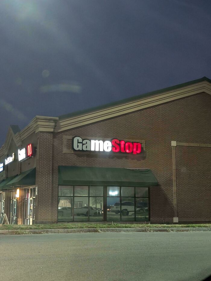 LED sign displaying GameStop logo on a brick building at night with illuminated storefront windows and parking area.