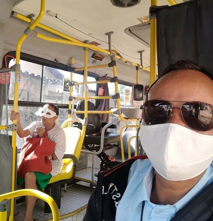 Man wearing mask and sunglasses taking selfie on bus, with passenger holding pole and wearing unusual eye mask inside.
