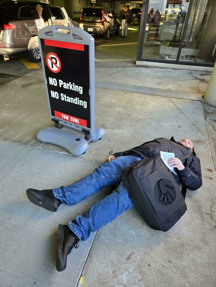 Man lying on the ground next to a no parking no standing sign in a humorous unhinged pics scenario.