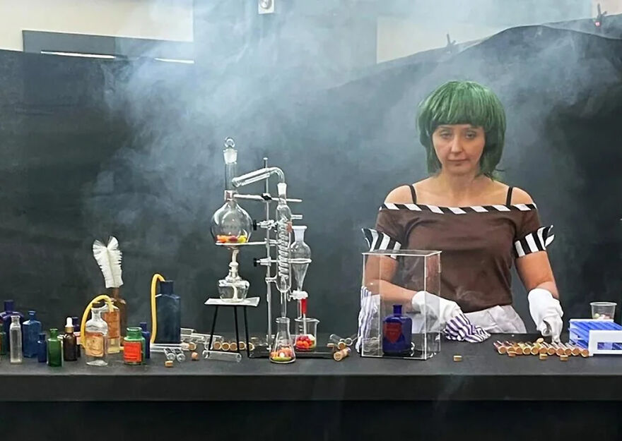 Woman with green wig and gloves performing a science experiment with glassware amid smoke, featuring 2024 pop culture moments.