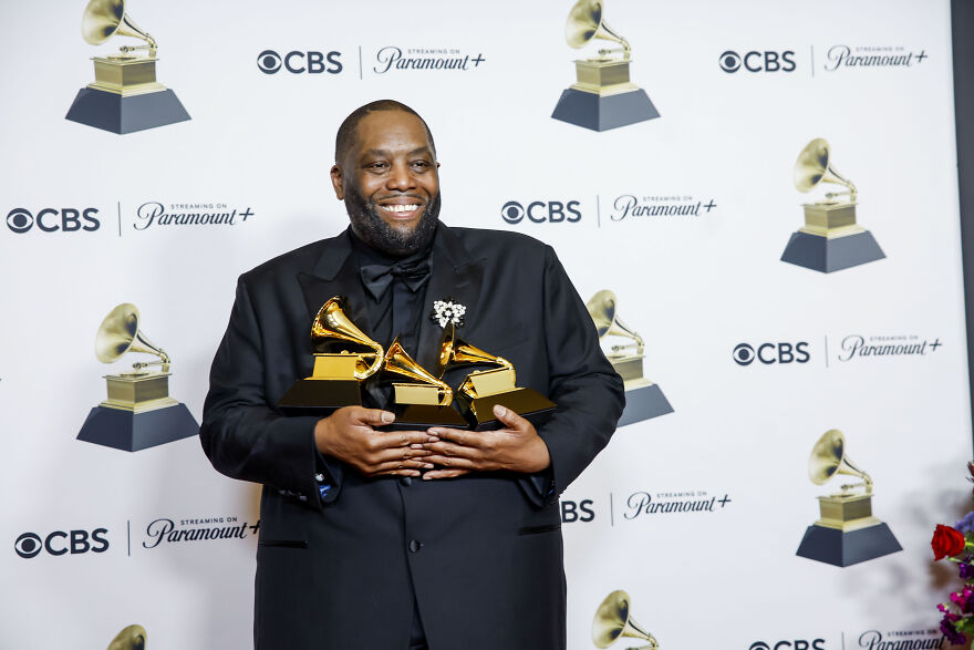 Man in black suit smiling and holding multiple Grammy awards at pop culture event with CBS and Paramount+ logos.