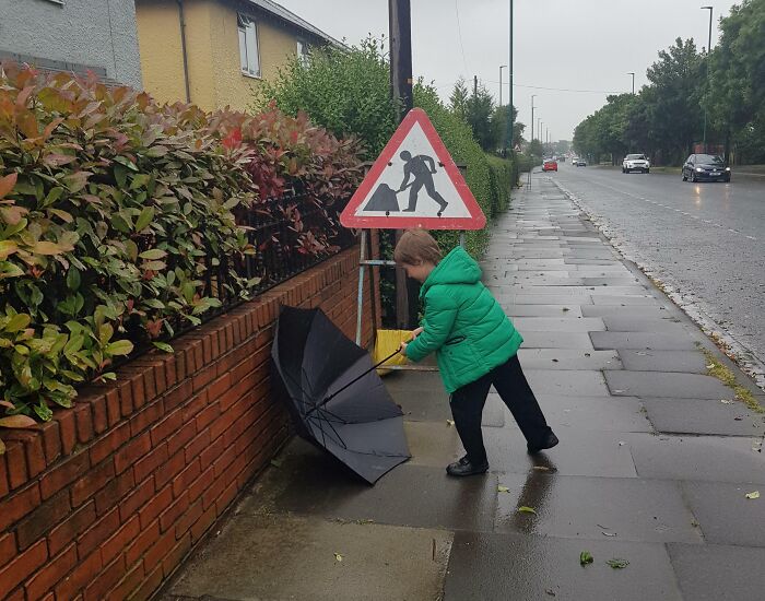 Child in green jacket mimics road work sign by holding a shovel and umbrella on a wet sidewalk outdoors.