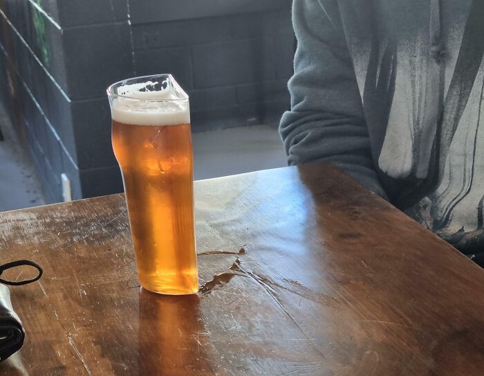 Tall glass of beer on a wooden table with a person nearby, showing unhinged pics of people doing exactly what they were asked to do.