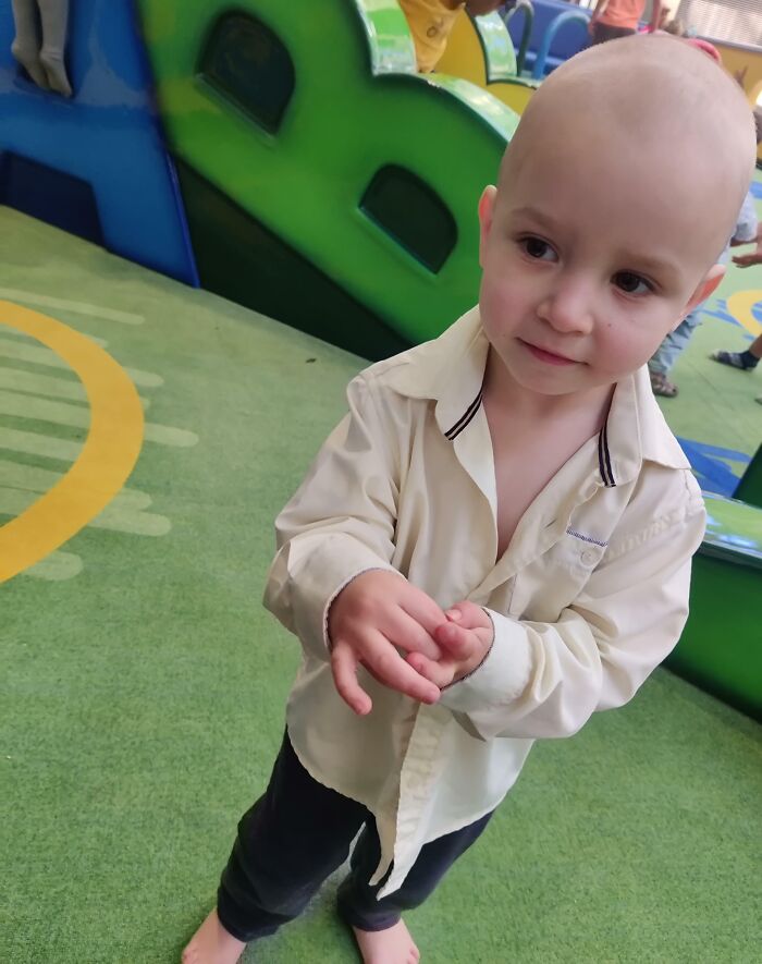 Young child with a bald head wearing a light shirt, standing barefoot on play area carpet, showing a hair accident moment.