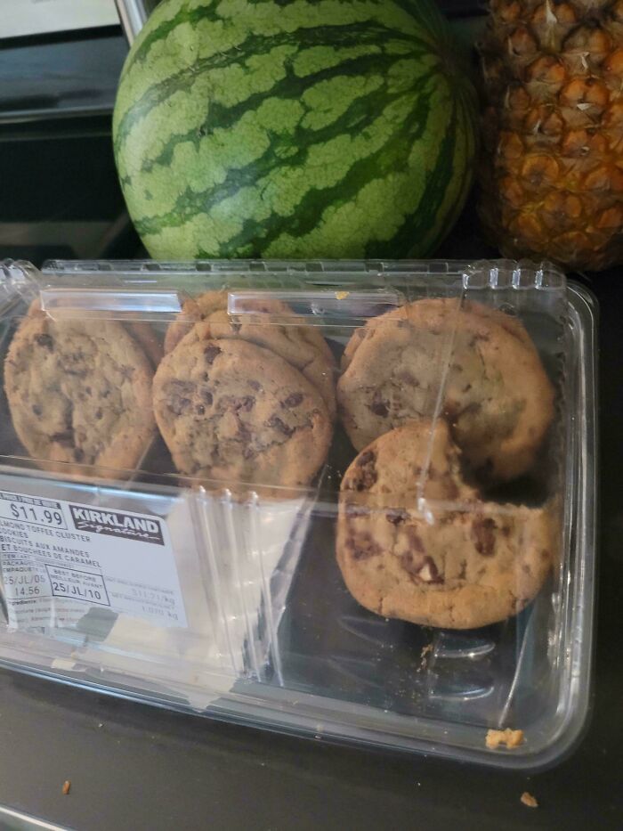 Plastic container of cookies with one cookie missing a large bite, placed near a watermelon and pineapple, showing family frustration.