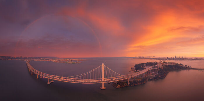 Panoramic view of a large suspension bridge at sunset with city skyline and rainbow in the background.