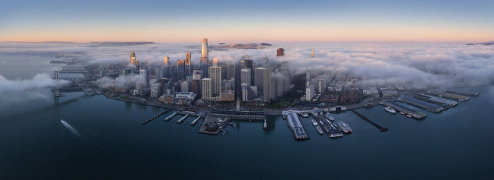 A stunning panoramic masterpiece of a fog-covered city skyline with piers and boats at sunrise, showcasing the Epson Pano Awards.