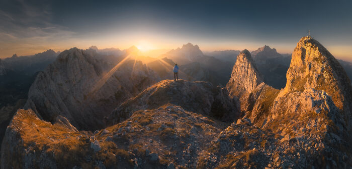 Panoramic masterpiece of a person standing on rugged mountain peak during golden sunset with expansive alpine landscape view.