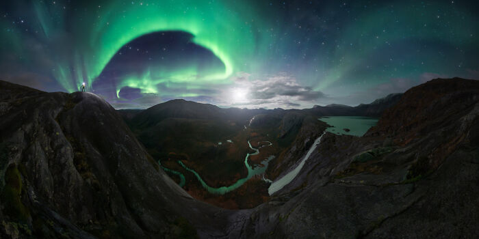 Panoramic landscape of a river valley at night with vibrant northern lights arching over rugged mountains.