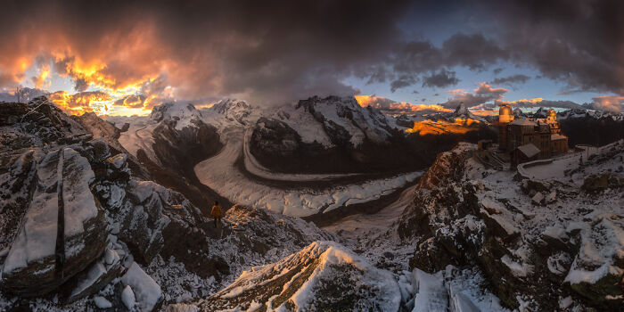 Panoramic mountain landscape with snowy peaks, glowing sunset sky, and a person standing on rocky terrain at dusk.