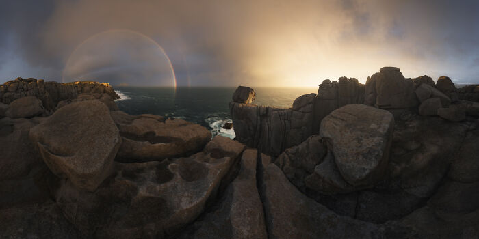 Panoramic view of rocky coastline at sunset with a faint rainbow, showcasing a panoramic masterpiece from Epson Pano Awards.