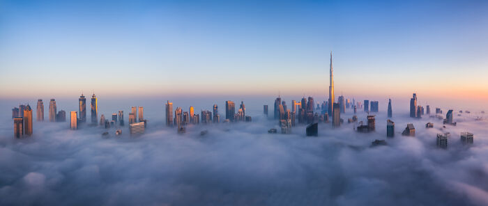 Panoramic view of a city skyline with skyscrapers rising above a dense layer of clouds during sunrise.