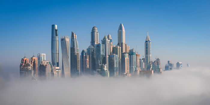 Panoramic cityscape with modern skyscrapers rising above thick fog under a clear blue sky, showcasing architectural beauty.