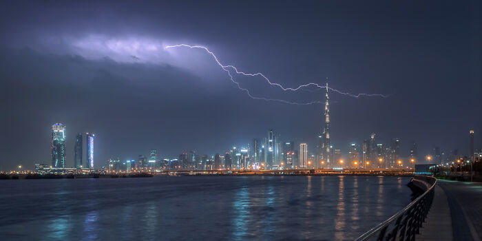 Panoramic night skyline with lightning striking over a cityscape featuring tall skyscrapers and reflecting on calm water.