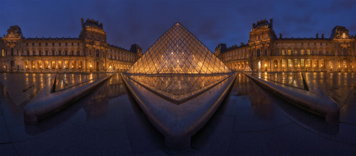 Panoramic nighttime view of the illuminated Louvre Pyramid and surrounding historic buildings in Paris.