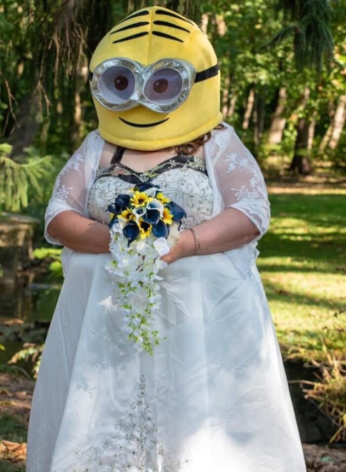 Bride in a wedding dress with lace sleeves holding a bouquet and wearing a large cartoon character helmet outdoors.
