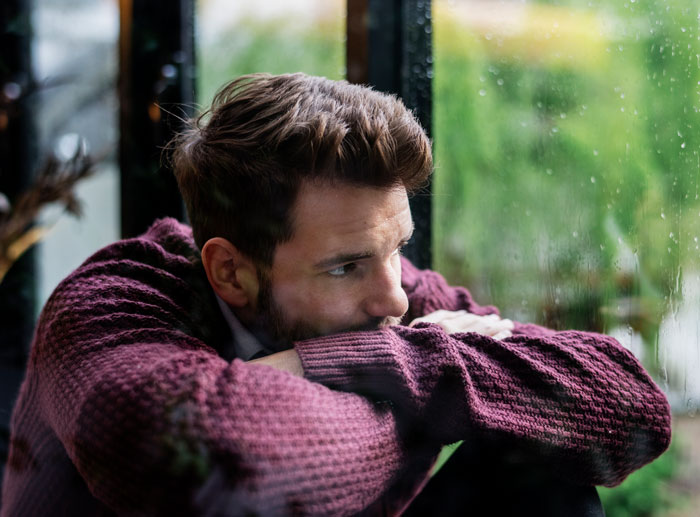 Man sitting by a rainy window looking sad and hurt after his wife asks to cancel their anniversary trip surprise. Man sitting by a rainy window looking sad and hurt after his wife asks to cancel their anniversary trip surprise.
