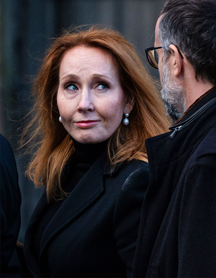 J.K. Rowling in a black coat with pearl earrings, engaging in conversation with a man wearing glasses and a dark jacket. J.K. Rowling in a black coat with pearl earrings, engaging in conversation with a man wearing glasses and a dark jacket.