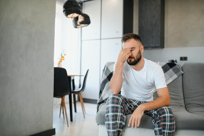 Man sitting on a couch at home, looking stressed and tired, while a woman does cardio with a toddler and vacuum nearby. Man sitting on a couch at home, looking stressed and tired, while a woman does cardio with a toddler and vacuum nearby.