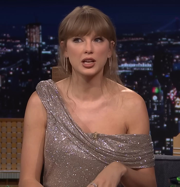 Taylor Swift wearing a sparkling one-shoulder dress, speaking during a nighttime talk show interview. Taylor Swift wearing a sparkling one-shoulder dress, speaking during a nighttime talk show interview.