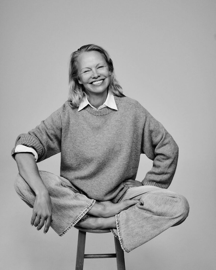 Pamela Anderson smiling seated cross-legged on a stool wearing a sweater and jeans showing confidence and reinvention. Pamela Anderson smiling seated cross-legged on a stool wearing a sweater and jeans showing confidence and reinvention.