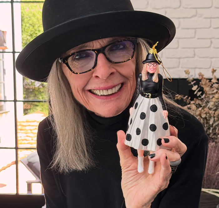 Diane Keaton smiling, wearing a black hat and glasses, holding a doll dressed in a black and white polka dot outfit. Diane Keaton smiling, wearing a black hat and glasses, holding a doll dressed in a black and white polka dot outfit.