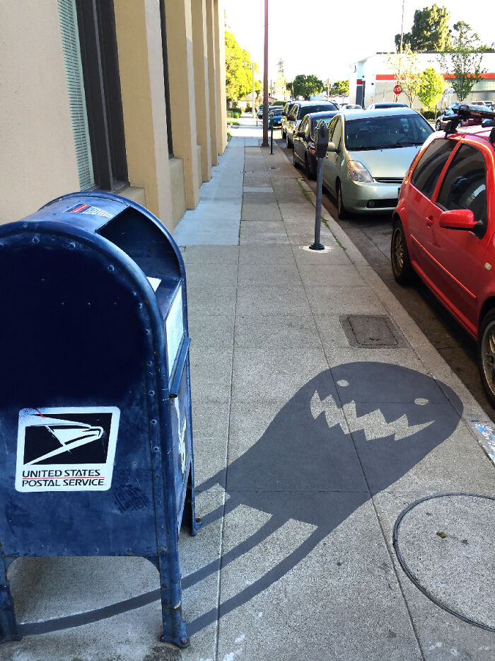 Shadow art installation showing a mailbox casting a playful monster-shaped shadow on the sidewalk at sunset.