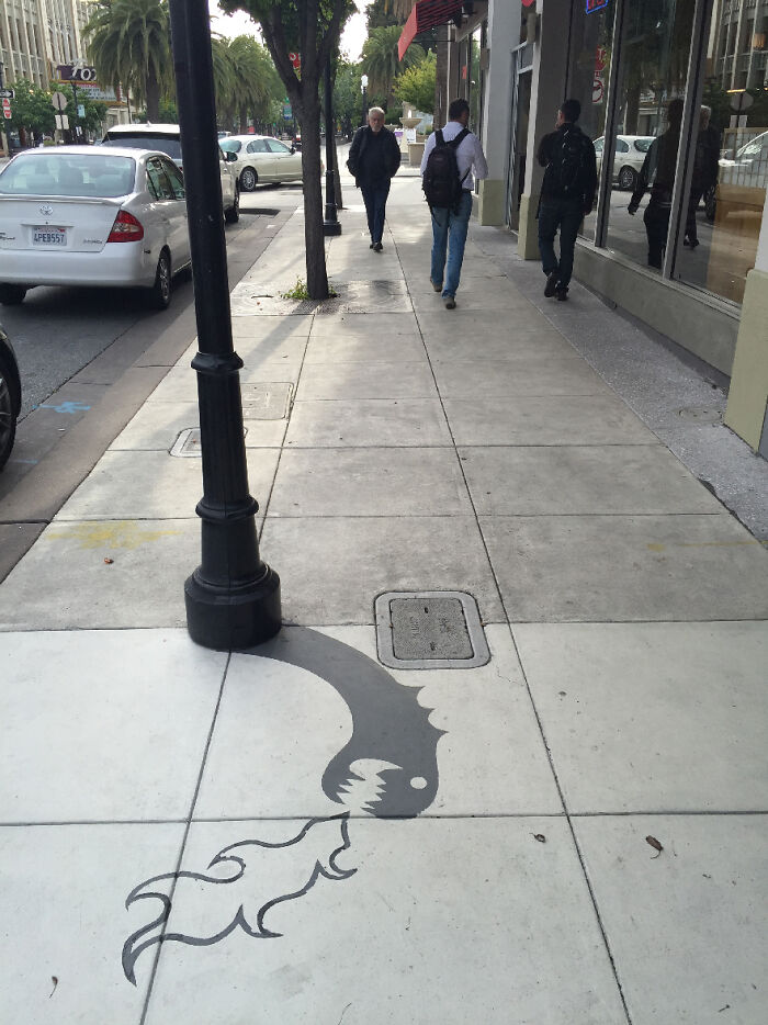 Street lamp shadow art installation on sidewalk creatively transforms shadow into a dragon breathing fire in clever shadow art.