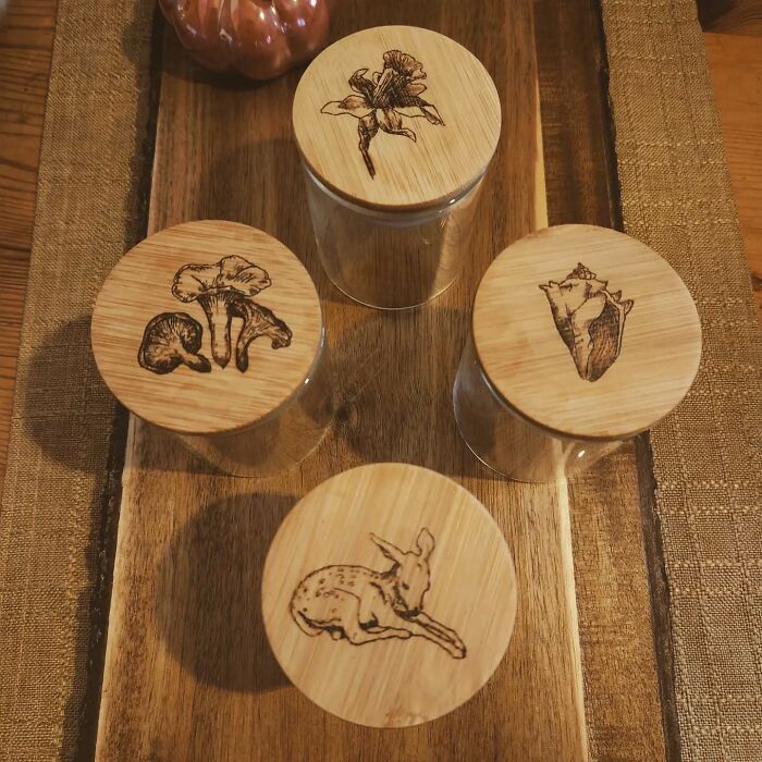 Wood-burned artwork by Canadian artist Kat Slota featuring detailed nature designs on wooden jar lids.