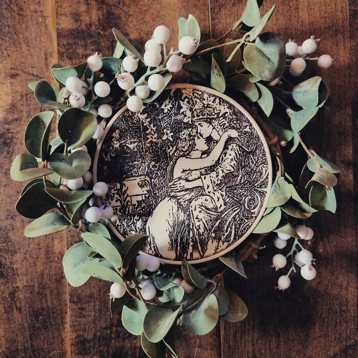 Wood-burned artwork by Canadian artist Kat Slota depicting an intimate scene, surrounded by green leaves and white berries.