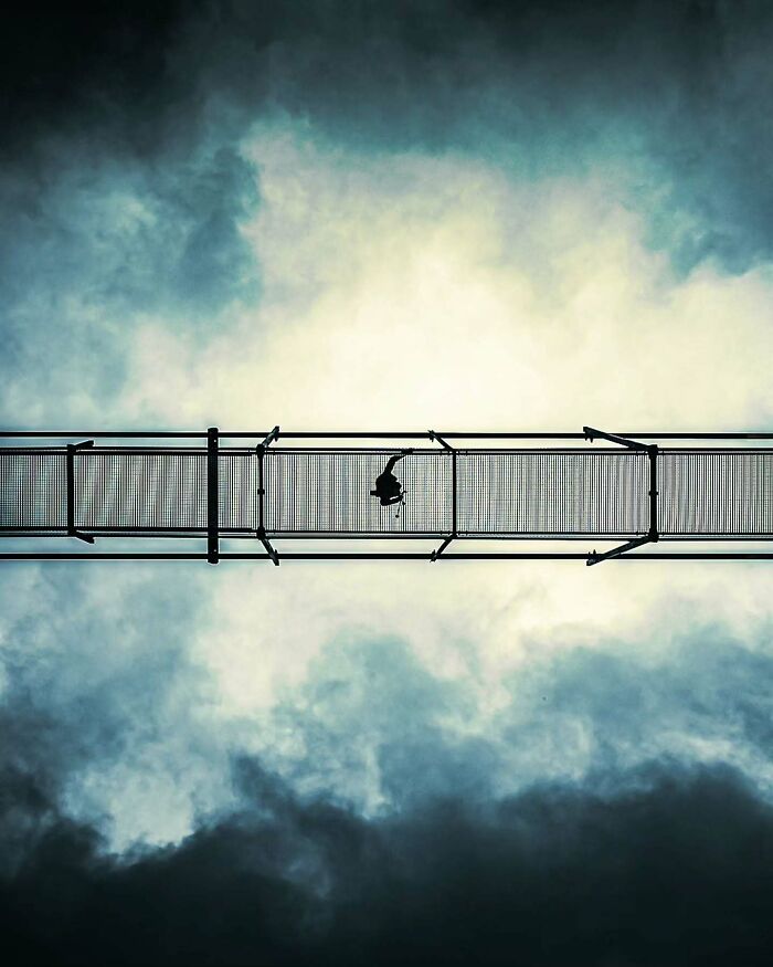 Minimalist architectural vision shown in a narrow bridge with a lone figure above dramatic cloudy sky.