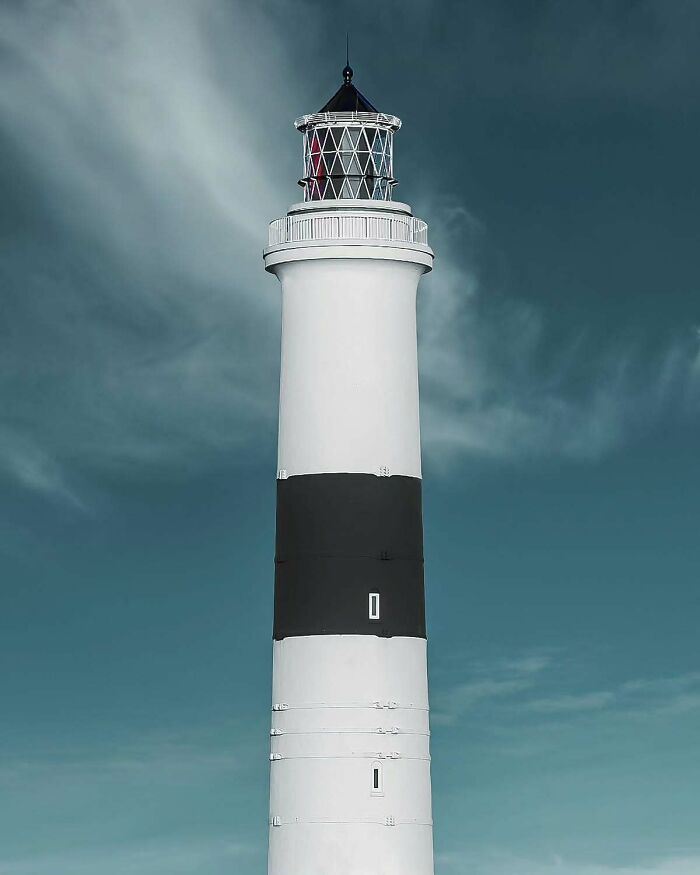 Minimalist architectural design of a white lighthouse with a black band under a moody cloudy sky.
