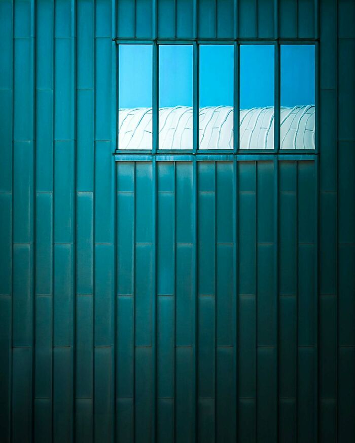 Minimalist architectural design showing teal metal panels with a rectangular window reflecting a white curved structure and sky.