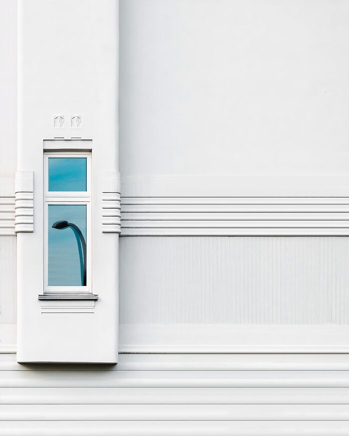 Minimalist architectural facade with clean white lines and a single window reflecting a streetlamp in daylight.