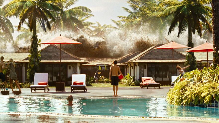 A huge ocean wave crashes behind tropical resort bungalows as people swim and relax by the pool in a true story movie scene.