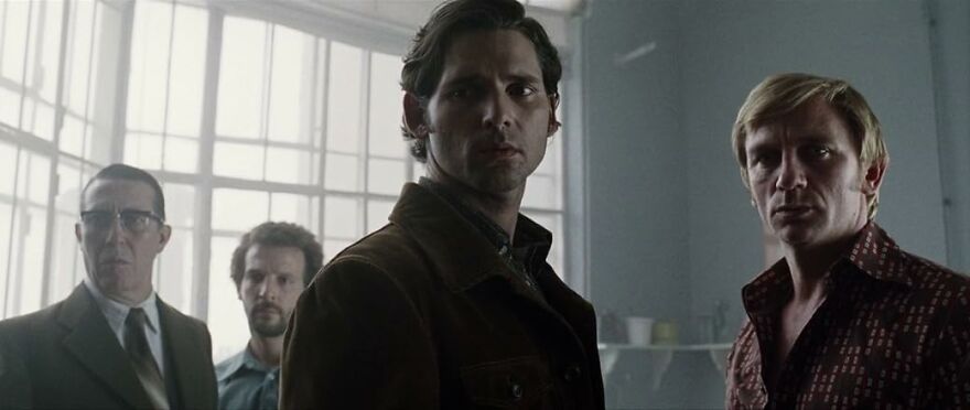 Four men with serious expressions stand in a dimly lit room, depicting a scene from movies based on true stories.