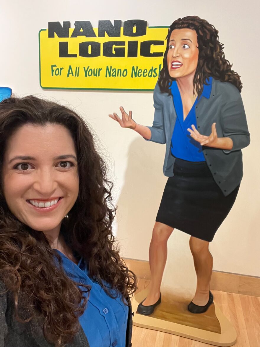 Woman with curly hair smiling in front of a colorful cartoon figure and a bright yellow sign about nano needs Woman with curly hair smiling in front of a colorful cartoon figure and a bright yellow sign about nano needs