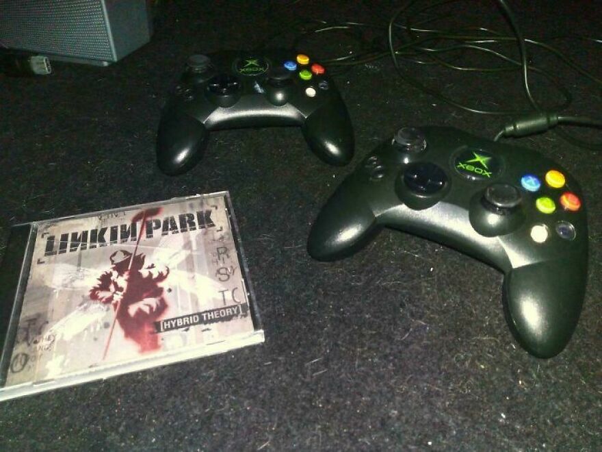 Two original Xbox controllers and a Linkin Park Hybrid Theory CD on a dark surface, evoking nostalgic memories.