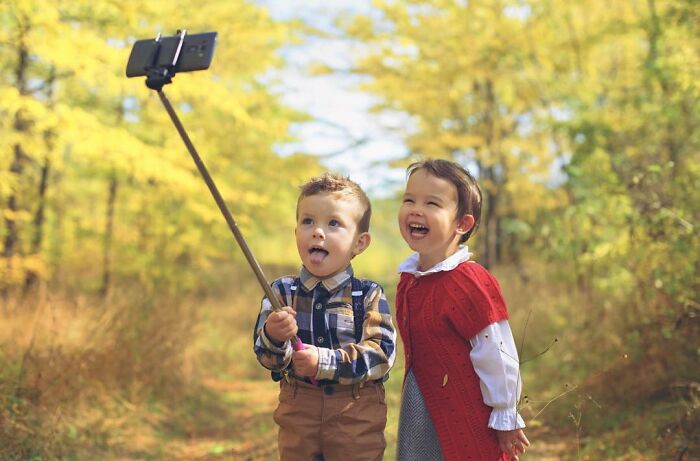 Two children smiling and taking a selfie with a smartphone on a selfie stick in a sunny forest, childhood photography.