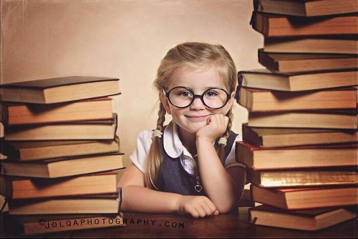 Young girl with glasses smiling between stacks of books, captured in a beautiful photo of childhood around the world.