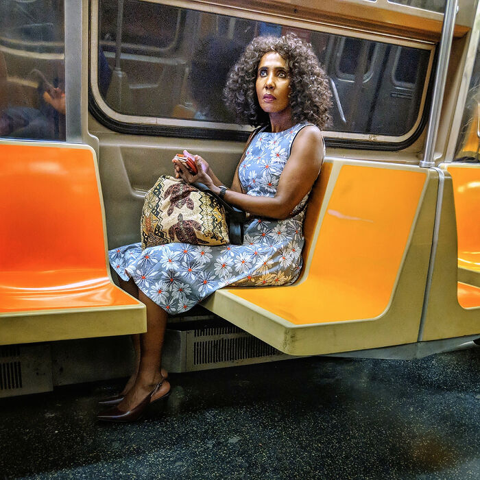 Woman in floral dress sitting on orange NYC subway seat, captured in a captivating street portrait underground.