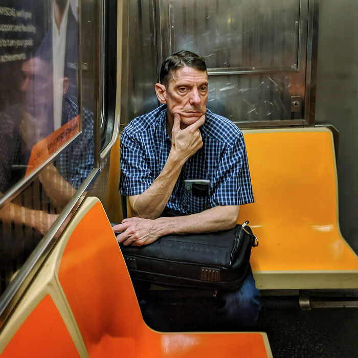 Middle-aged man in a blue checkered shirt sitting thoughtfully on an orange subway seat in NYC underground street portrait.