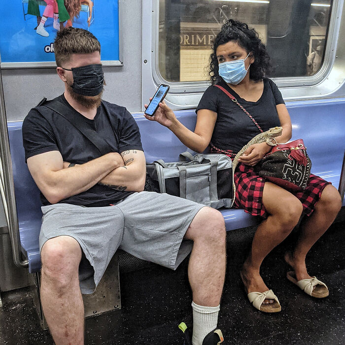 Two masked passengers sitting on NYC subway seats, one showing phone screen to the other, capturing life underground street portraits.